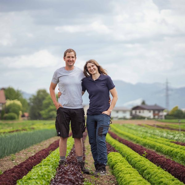 Ein Paar steht zwischen Gemüse- und Salatreihen auf einem Feld, im Hintergrund Berge und Häuser.