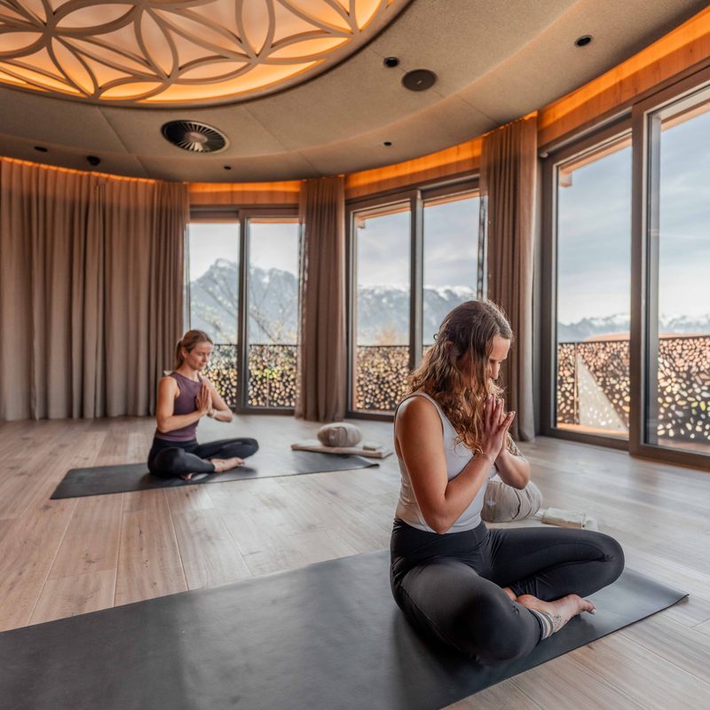 Zwei Frauen meditieren auf Yogamatten in einem Wellnessraum mit großer Fensterfront und Blick auf Berge.