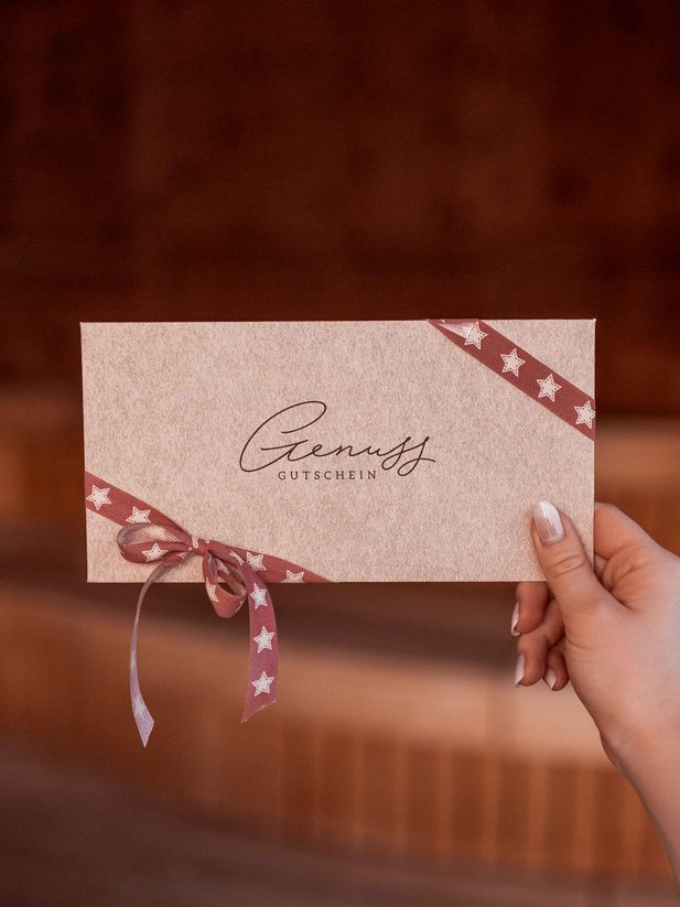 Hand holding an indulgence gift voucher with ribbon in front of a wooden background.