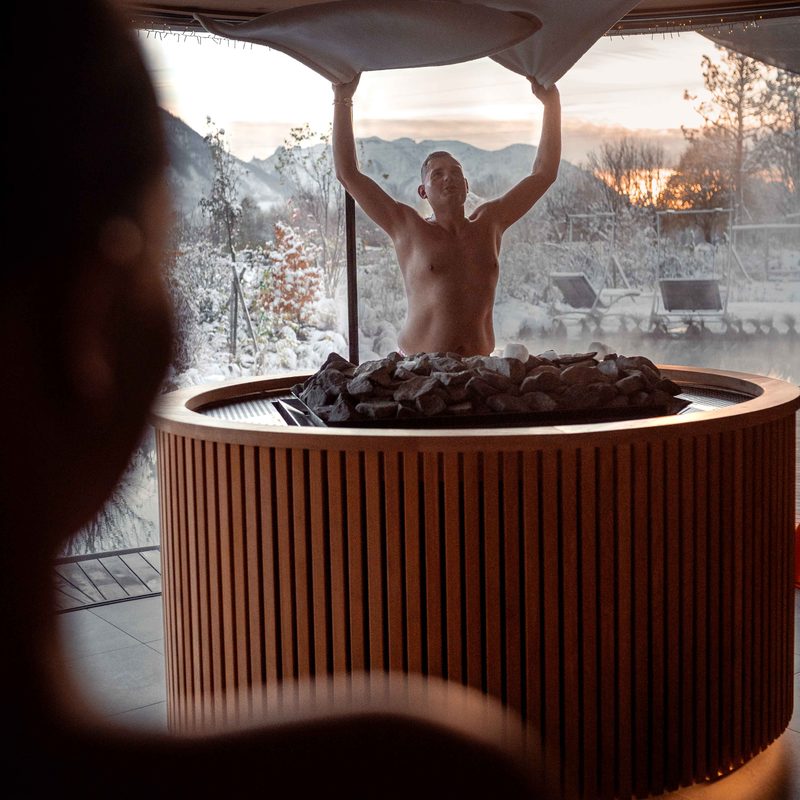 Guest doing a sauna infusion, waving a towel in front of a panoramic window with snowy mountain landscape.