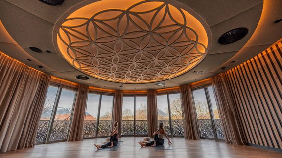 Two guests do yoga on mats in a wellness room with large windows and a round ceiling light.