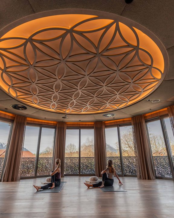 Zwei Gäste machen Yoga auf Matten in einem Wellnessraum mit großen Fenstern und runder Deckenleuchte.