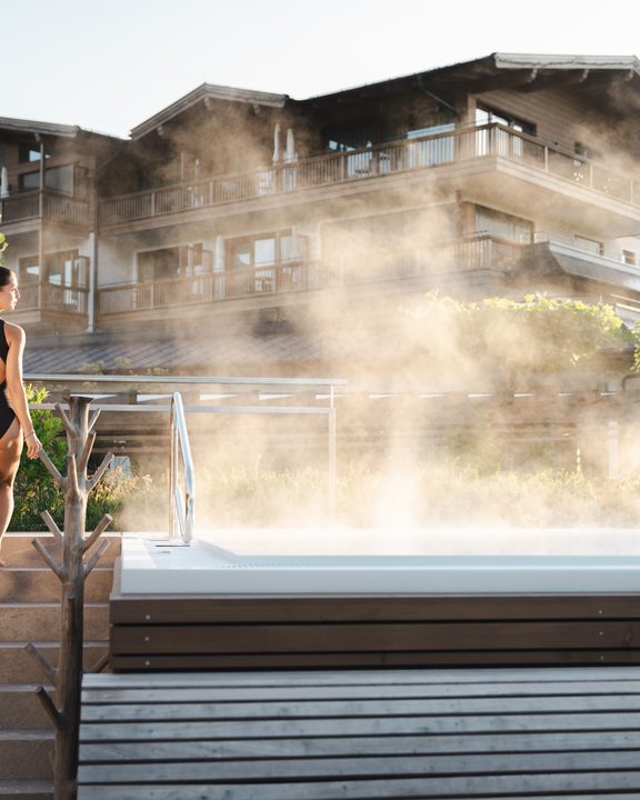 Frau im Badeanzug am dampfenden Außenpool vor einem Hotelgebäude.