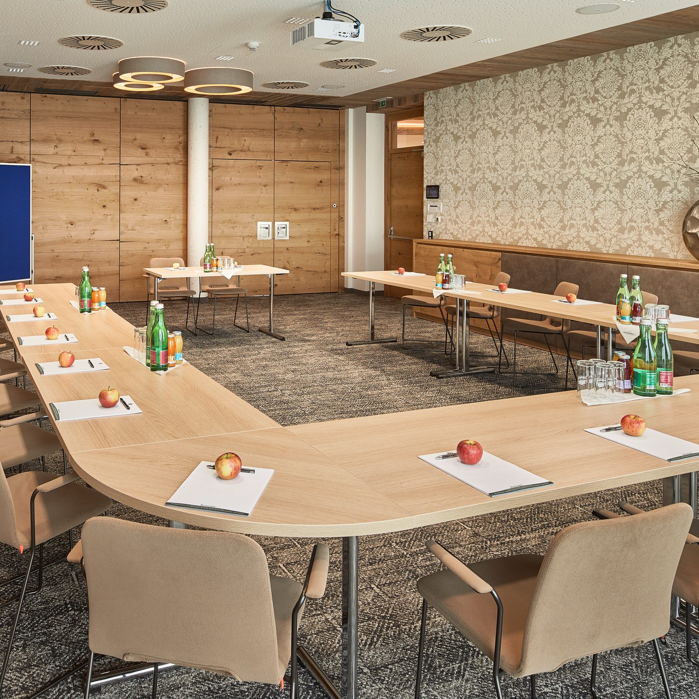 Meeting room with U-shaped table setup, presentation boards and ceiling projector, water bottles and apples on tables.