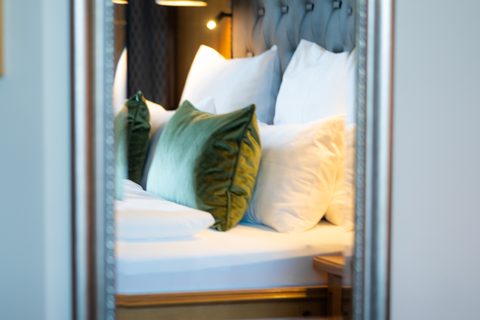 Gemachtes Hotelbett mit gepolstertem Kopfteil, weißen Kissen und grünen Zierkissen, im Spiegel aufgenommen.