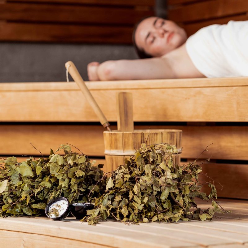 Woman lying on a wooden sauna bench with an infusion bucket, ladle and birch twigs in the foreground.