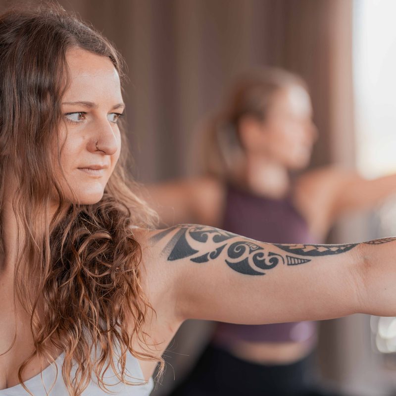 Frau macht Yoga-Pose mit ausgestrecktem Arm, im Hintergrund trainiert eine weitere Person.