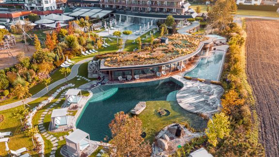 Drone view of a hotel wellness area with a natural pool, loungers, cabanas and landscaped garden in autumn.
