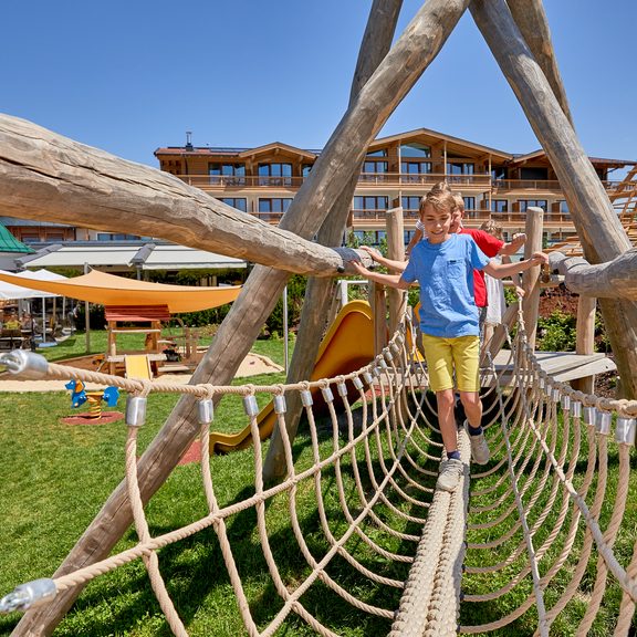 Zwei Kinder balancieren über eine Seilbrücke auf einem Spielplatz vor einem Hotel.