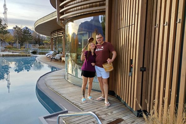 Two guests with sauna ladle and infusion bucket on a wooden deck by a wellness pool in front of a curved glass facade.