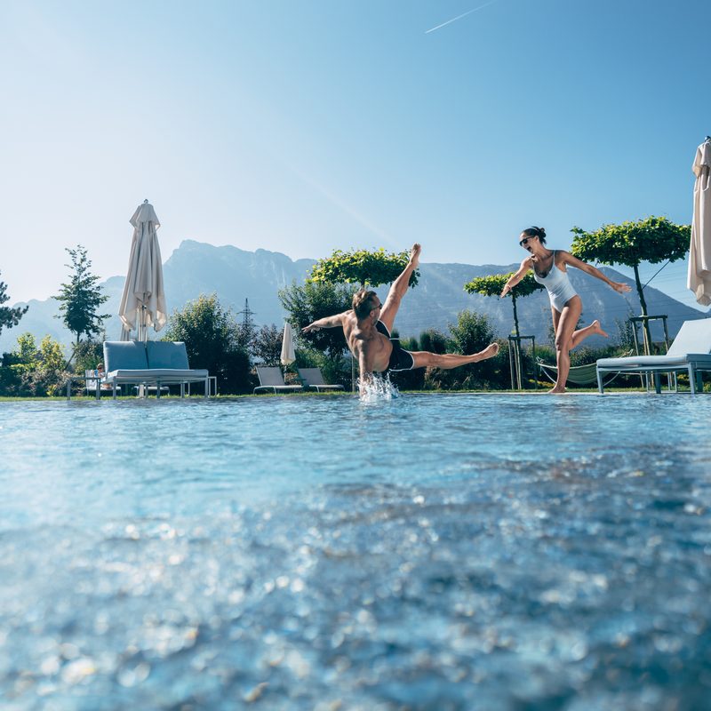 Ein Paar am Außenpool, während der Mann ins Wasser springt, mit Liegen, Sonnenschirmen und Bergkulisse.