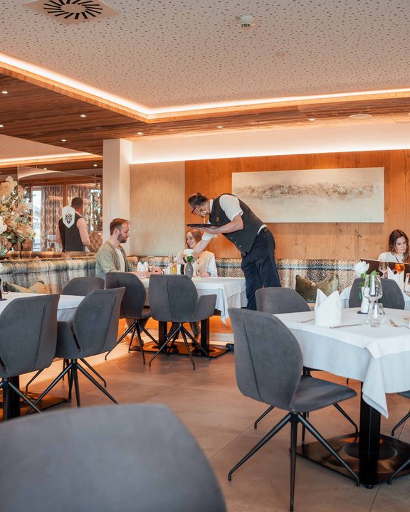 Innenansicht eines Hotelrestaurants mit gedeckten Tischen, Gästen und einem Kellner beim Servieren.