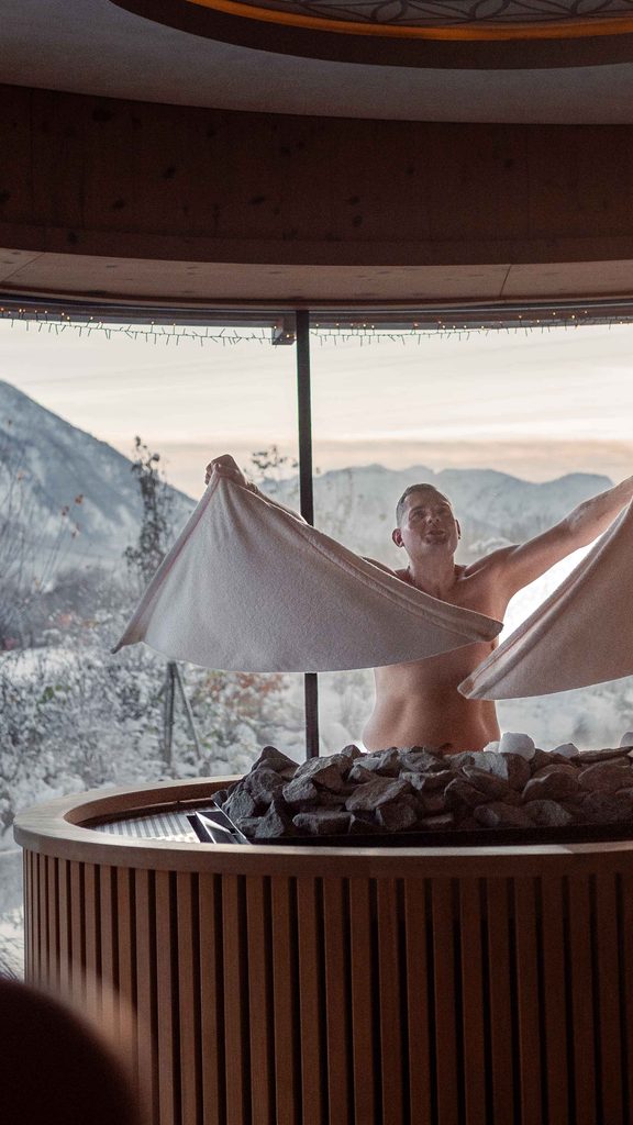 Saunagast schwingt ein Handtuch beim Aufguss vor Panoramafenster mit Blick auf verschneite Berge.