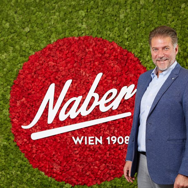 Mann steht vor grüner Mooswand mit rotem Kreis und Schriftzug „Naber – Wien 1908“.