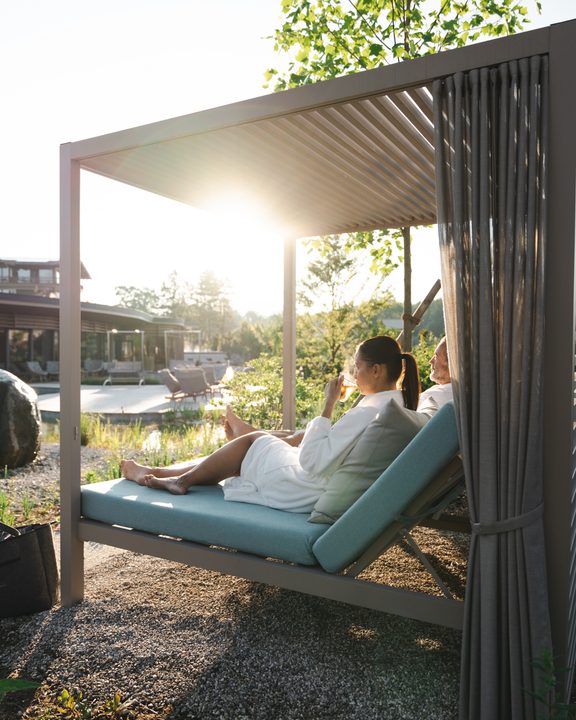 Zwei Gäste im Bademantel entspannen auf einem Outdoor-Daybed mit Vorhängen im Wellness-Außenbereich.