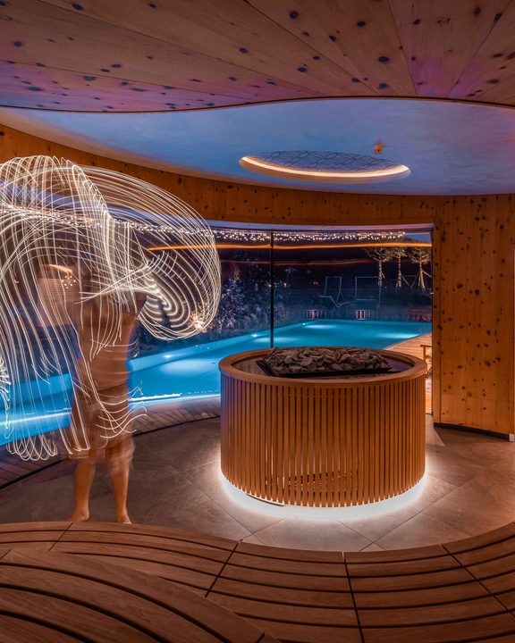 Guest in spa area by a lit pool with long-exposure light trails and a sauna in the background.