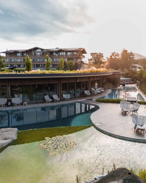 Naturpool mit Holzdeck und Liegestühlen vor moderner Hotelanlage im Abendlicht.