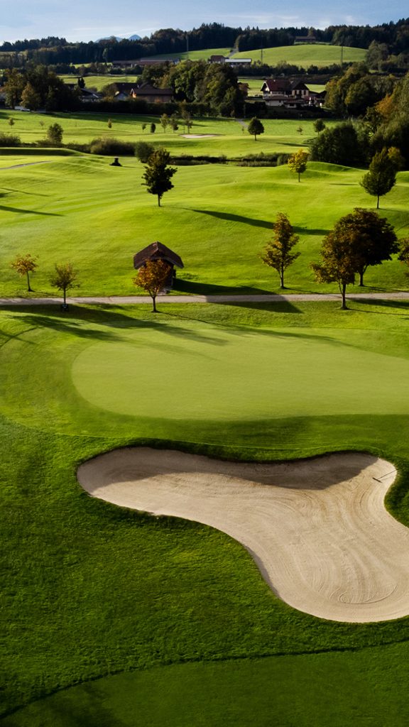 Golf green with a sand bunker in rolling countryside with trees and fields.