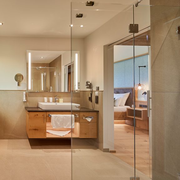 Modern hotel bathroom with wooden vanity, mirror and glass shower, view through the doorway into the bedroom.