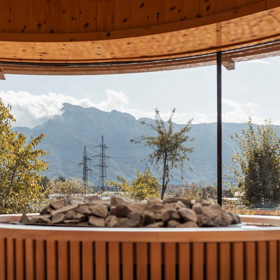 Sauna-Innenraum mit Saunasteinen vor Panoramafenster und Blick auf Berge und Bäume.