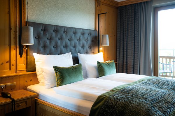 Doppelbett mit gepolstertem Kopfteil, grünen Kissen und Balkonfenster in einem Hotelzimmer.