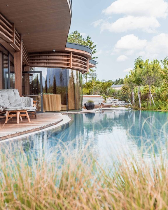 Holzterrasse mit Loungesesseln am Naturpool neben einem modernen Rundbau, umgeben von Gartenpflanzen.