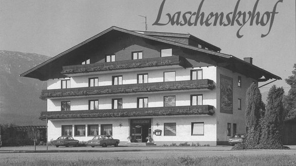 Black-and-white exterior view of the Laschenskyhof with balcony façade, parked cars and a mountain backdrop.