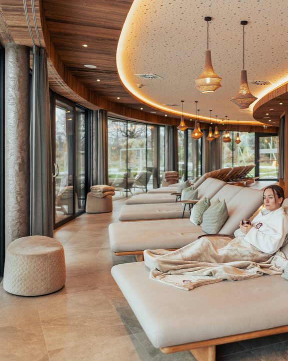 Woman relaxing on a daybed in a spa relaxation room by a large glass wall.