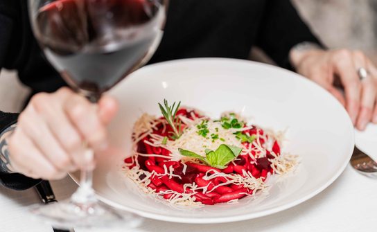Plate of red pasta with grated cheese and herbs next to a glass of red wine held by a guest.