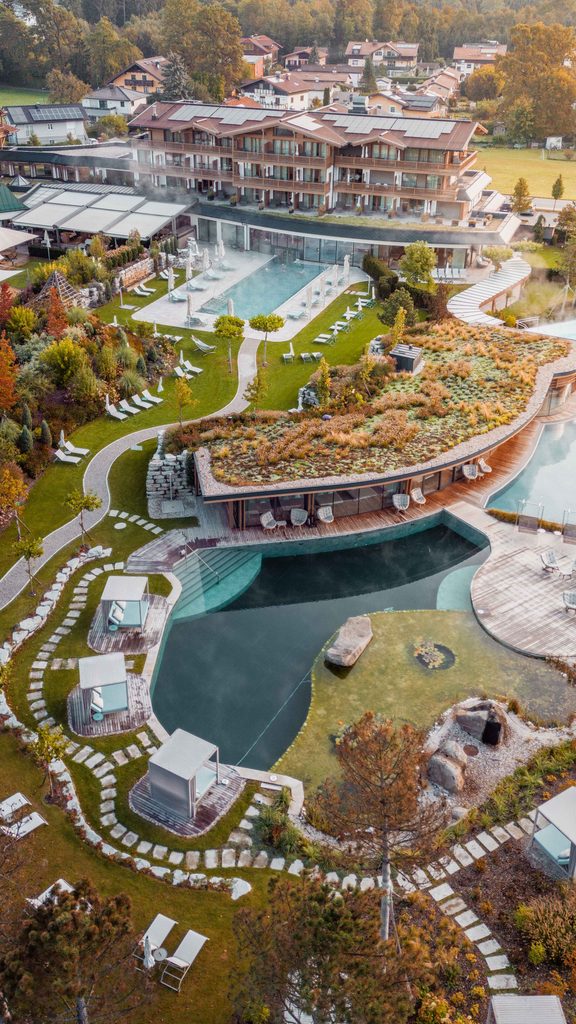 Luftaufnahme eines Hotels mit Wellnessgarten, Außenpools und Naturbadeteich in herbstlicher Landschaft.