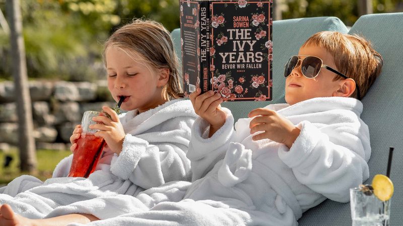 Zwei Kinder im Bademantel auf einer Outdoor-Liege, eines liest ein Buch, beide trinken Getränke im Garten.