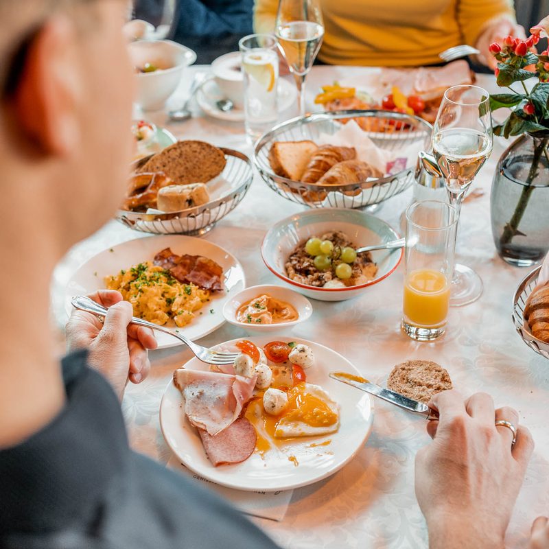 Zwei Gäste sitzen am Frühstückstisch mit Brot, Croissants, Aufschnitt, Obst, Orangensaft und Wasser.