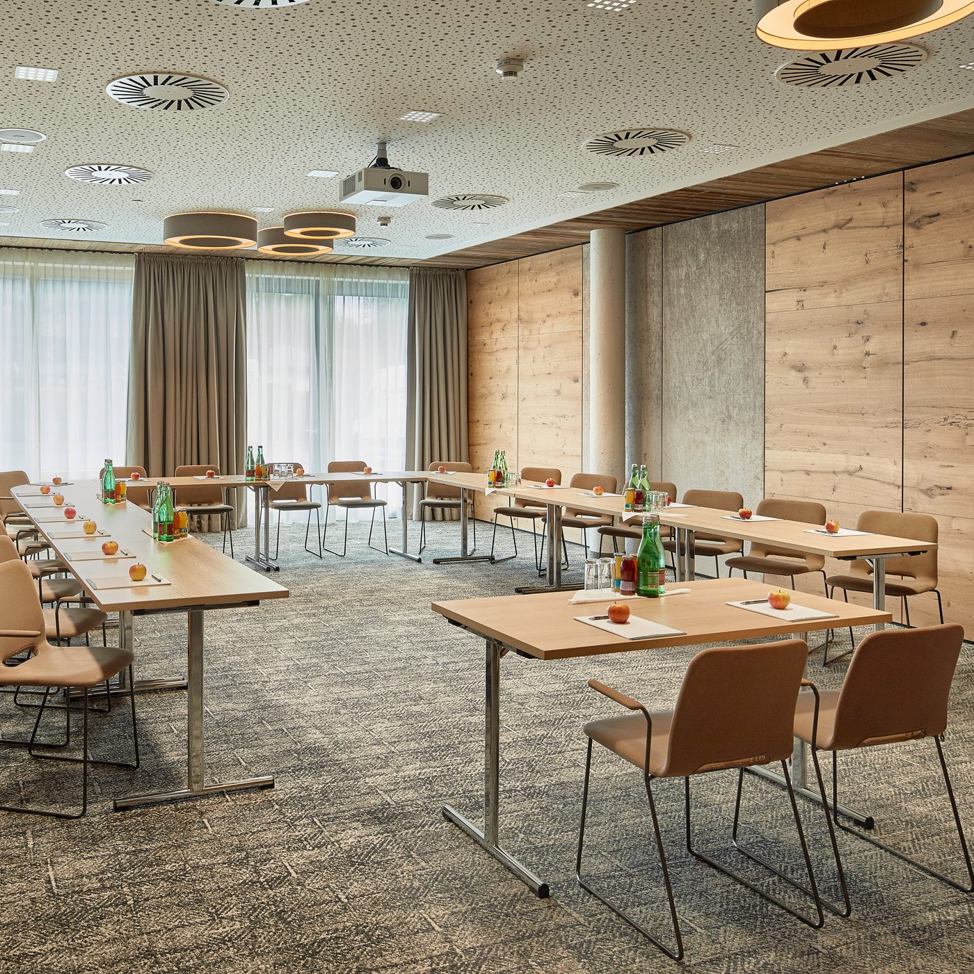 Seminar room with U-shaped seating, tables, water bottles and a ceiling projector in front of large windows.