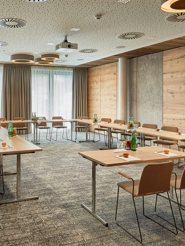 Seminar room with U-shaped seating, tables, water bottles and a ceiling projector in front of large windows.