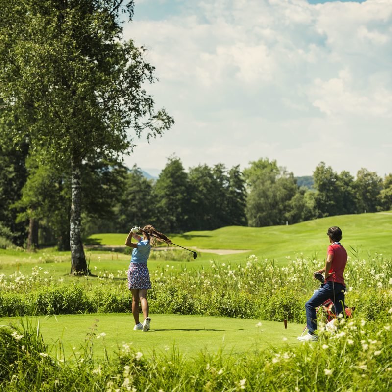 Two guests on a golf course at the tee, with golf club, golf bag and wide meadow landscape.
