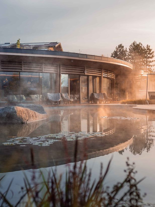 Moderner Wellnessbereich mit Naturpool, Liegen und aufsteigendem Dampf vor einer Glasfassade.