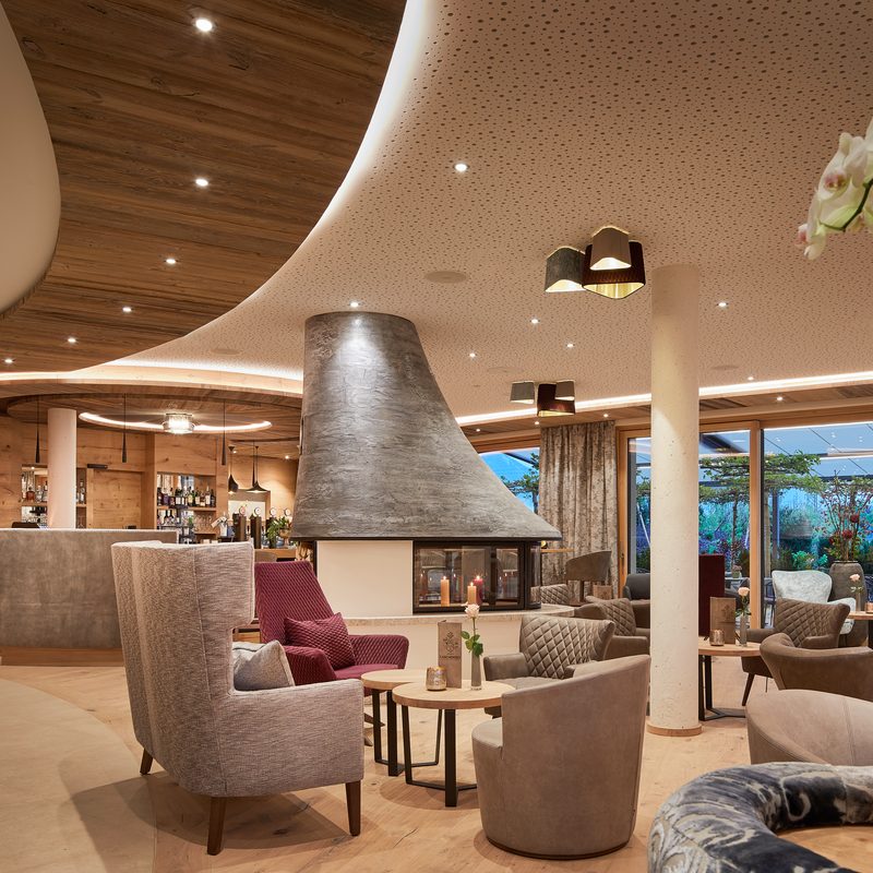 Hotel-Lounge mit Sesseln, Kamin und Barbereich vor einer Glasfront mit Terrassenzugang.