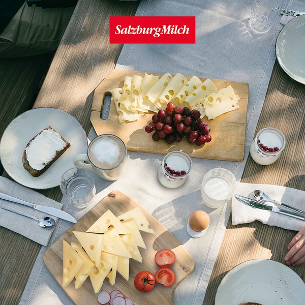Gedeckter Frühstückstisch mit Käse, Trauben, Kaffee, Joghurt, Brot, Ei und frischem Gemüse auf Holzbrettern.