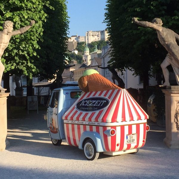 Rot-weiß gestreifter Eiswagen mit großem Eishorn zwischen Statuen am Parkeingang.