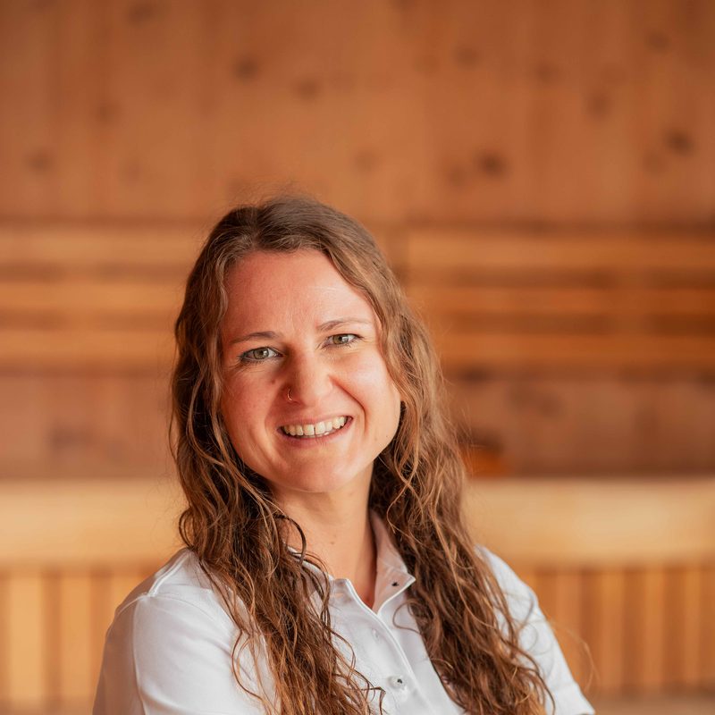 Smiling staff member in a spa area in front of a wooden sauna.