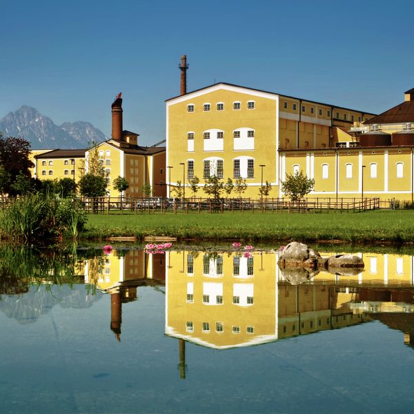 Gelber Gebäudekomplex spiegelt sich in einem Teich, im Hintergrund Berge unter blauem Himmel.