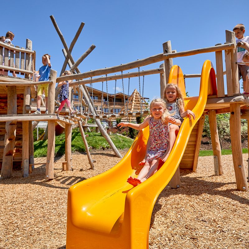 Kinder rutschen eine gelbe Doppelrutsche auf einem Holzspielplatz im Freien hinunter.