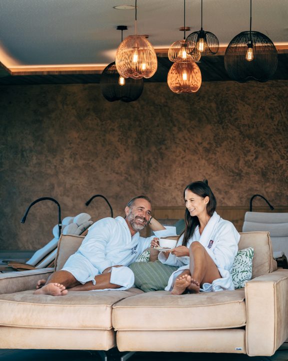 Couple in bathrobes relaxing on a sofa in a wellness lounge area, drinking tea.