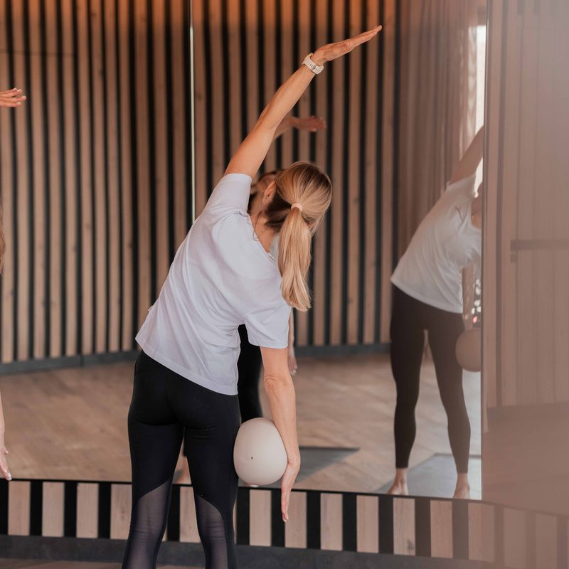 Two women do side stretching exercises in a fitness room with a mirror and yoga mats.