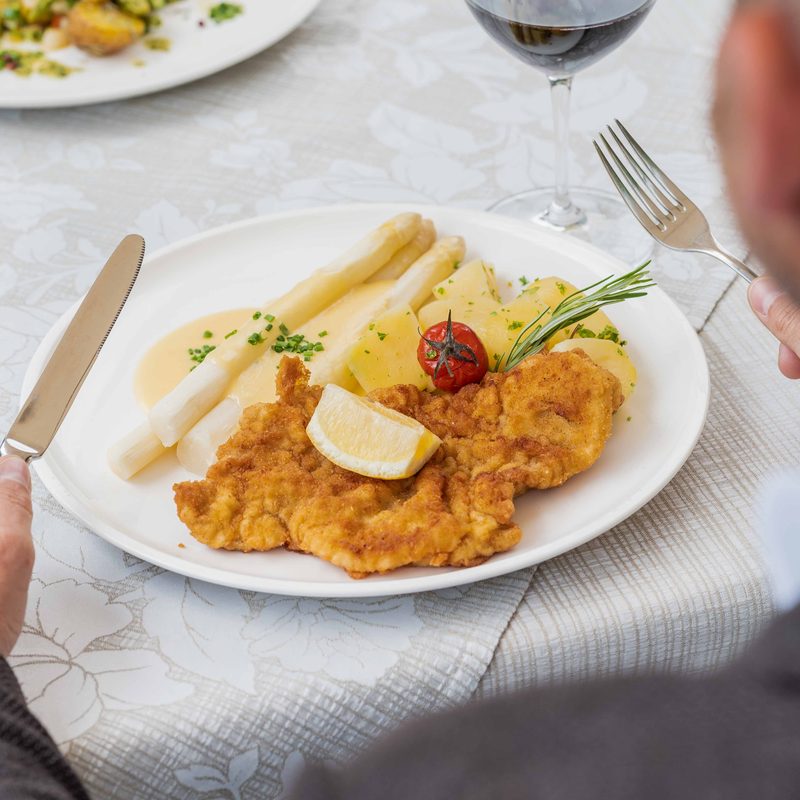 Paniertes Schnitzel mit weißem Spargel und Kartoffeln auf einem Teller, Gabel und Messer in den Händen eines Gastes.
