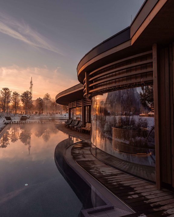 Dampfender Außenpool neben modernem Wellnessgebäude im Winter bei Sonnenaufgang.