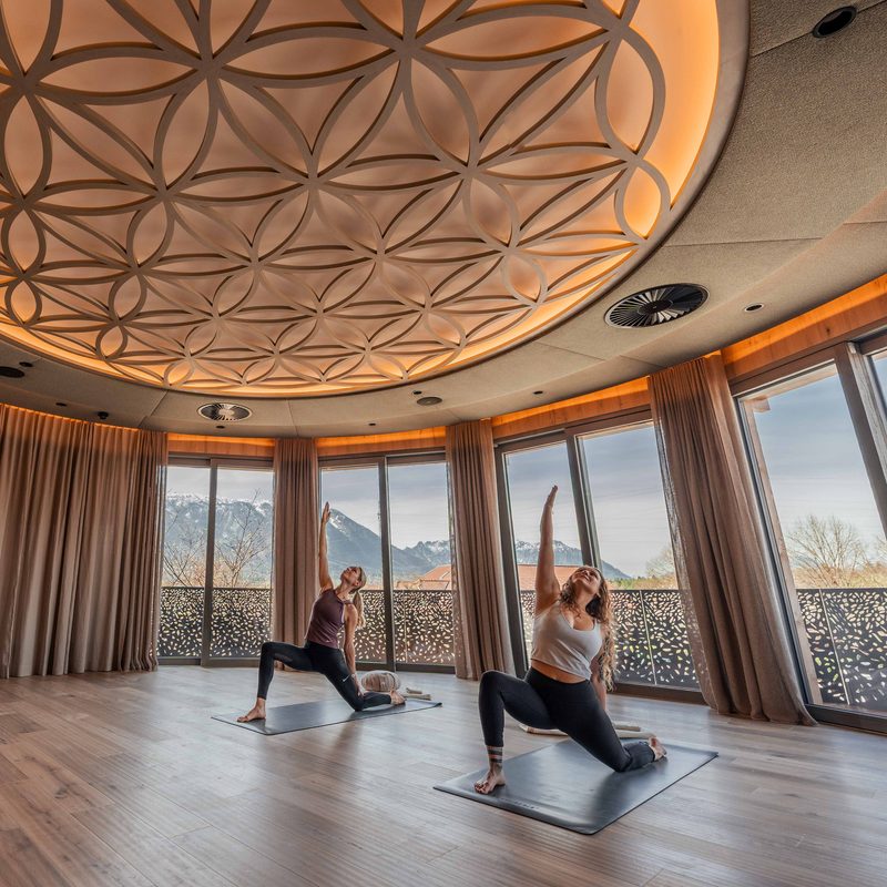 Zwei Personen machen Yoga auf Matten in einem Wellnessraum mit großen Fenstern und Bergblick.