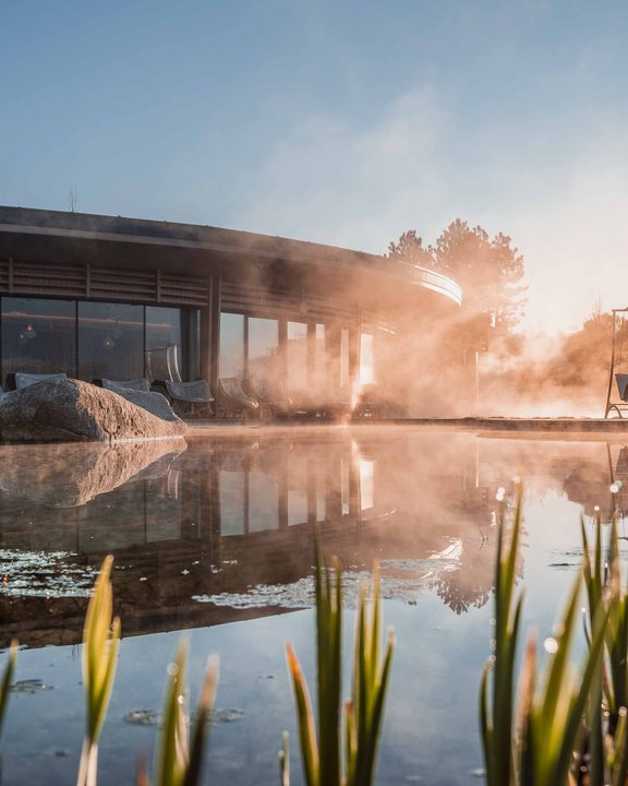 Dampf steigt über einem Außenpool vor einer Wellnessanlage in der Morgensonne auf.