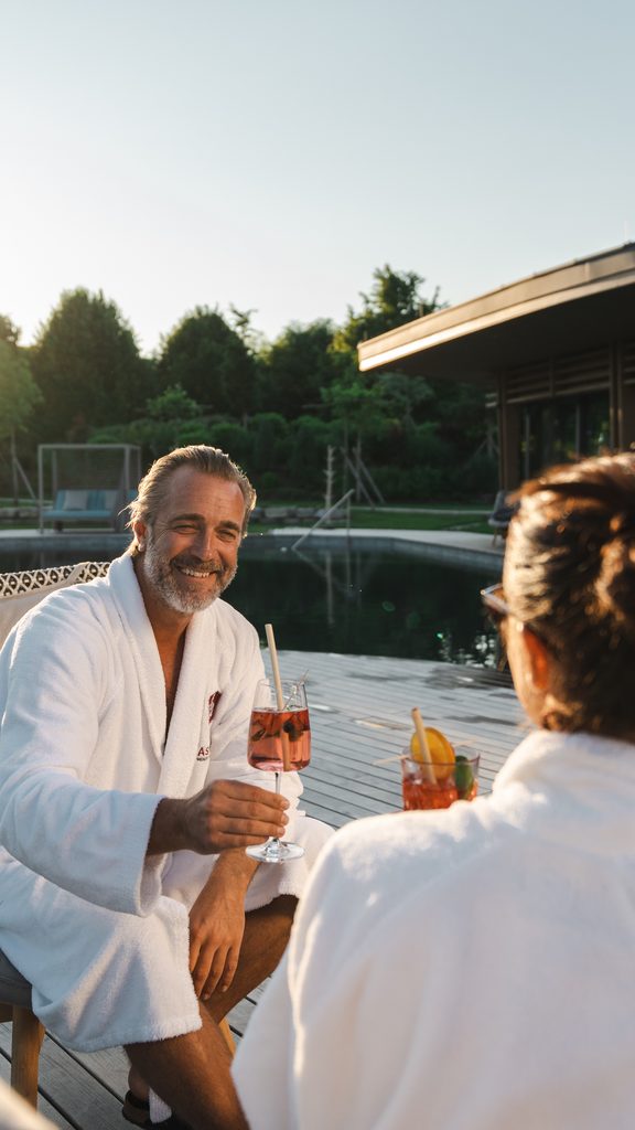 Zwei Gäste im Bademantel stoßen mit Drinks auf einer Poolterrasse bei Sonnenuntergang an.