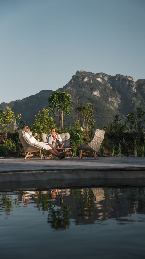 Zwei Gäste im Bademantel entspannen auf Liegestühlen am Pool mit Bergpanorama im Hintergrund.